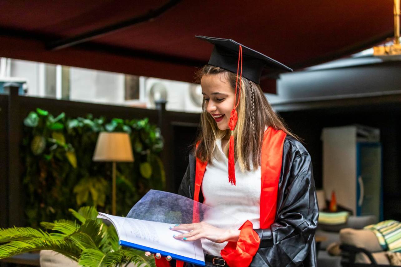 A smiling young woman in a black graduation cap and gown with red trim, looking down at an open book.